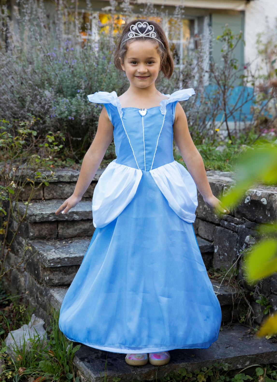 A young girl stands on stone steps outdoors, smiling while dressed in a blue princess costume from the Great Pretenders Boutique Princess Gown Collection with a tiara.