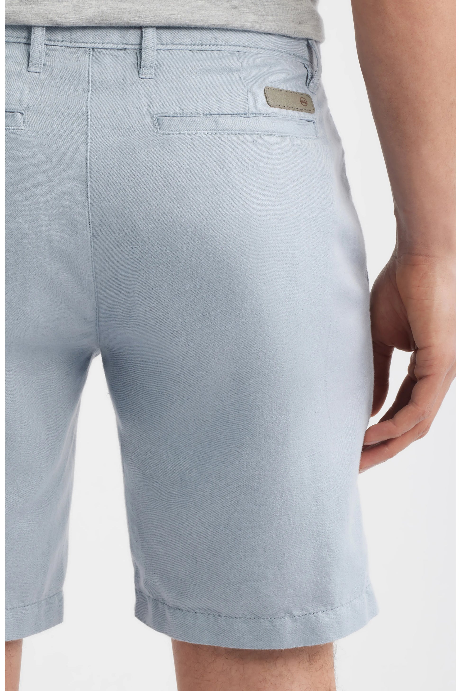 A person is shown from the back waist to above the knee wearing AG Jeans' Wanderer Shorts Linen in a light blue shade, featuring a slim cut, belt loops, and back welt pockets. One hand rests by their side, highlighting these warm weather shorts.