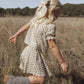A young child with blonde hair wears the Rylee & Cru Girls’ Liv Blouse, styled with boots, while walking through dry grass in a field with trees and hills in the background.