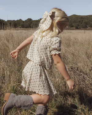 A young child with blonde hair wears the Rylee & Cru Girls’ Liv Blouse, styled with boots, while walking through dry grass in a field with trees and hills in the background.