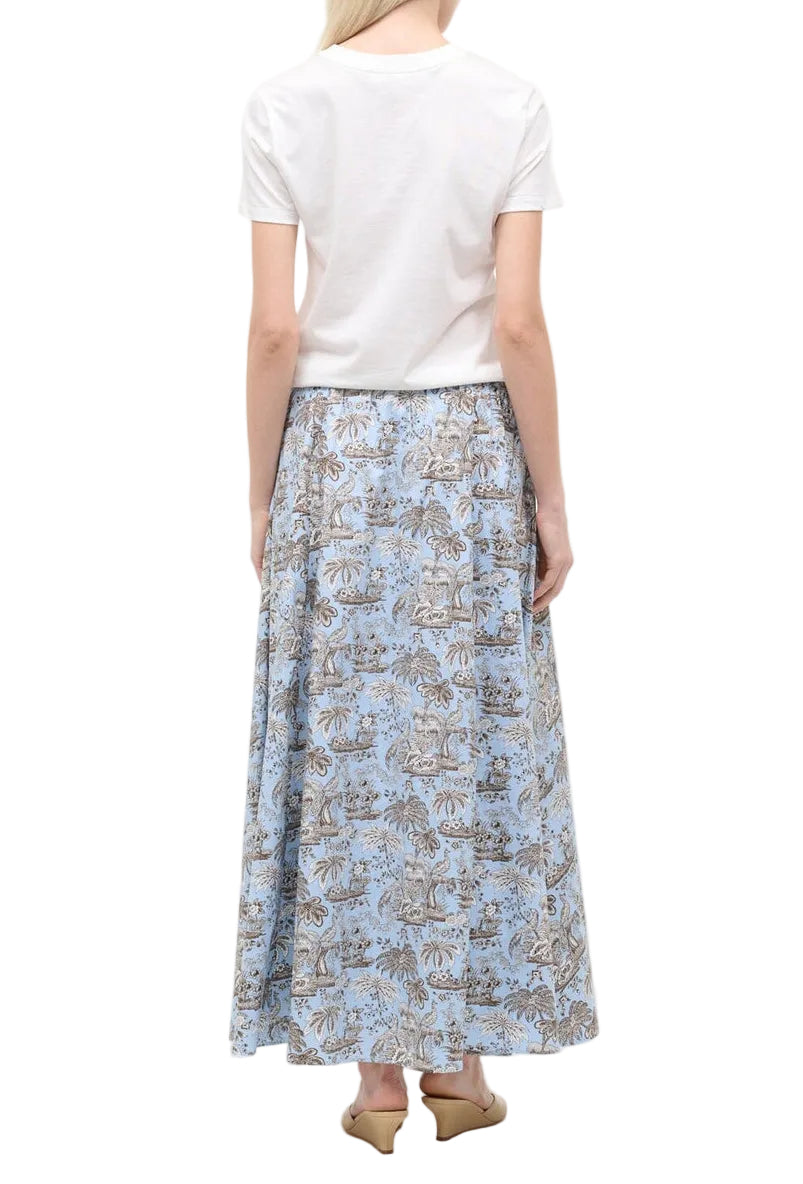 A woman facing away wears a white short-sleeve shirt, beige pointed-toe shoes, and the Staud Axel Skirt—a light blue floral pull-on maxi skirt.