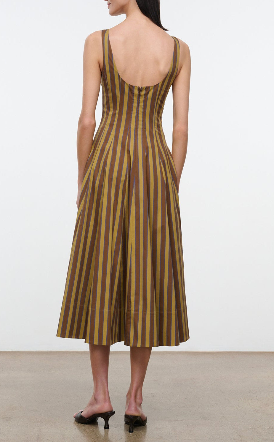 A woman stands on a plain floor facing away from the camera, wearing the Wells Midi Cotton Poplin Dress by Staud—a sleeveless design in brown and yellow stripes with a scoop back. This polished piece highlights Staud’s signature silhouette for an effortlessly refined look.