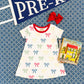 A Girls' Polly Play Dress by The Beaufort Bonnet Company, a red hair bow, a children's book, and a navy “PRE-K” pennant are displayed on a blue patterned surface.
