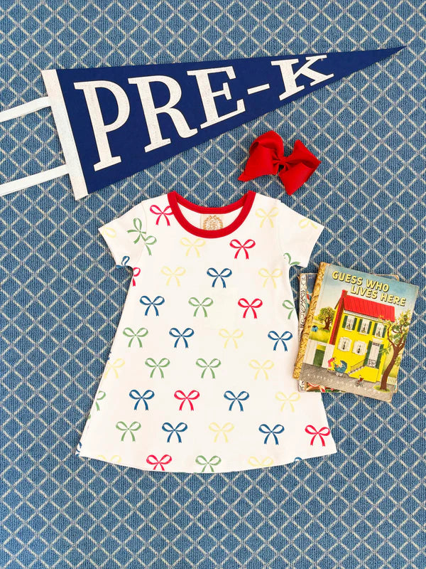 A Girls' Polly Play Dress by The Beaufort Bonnet Company, a red hair bow, a children's book, and a navy “PRE-K” pennant are displayed on a blue patterned surface.