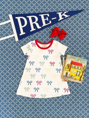 A Girls' Polly Play Dress by The Beaufort Bonnet Company, a red hair bow, a children's book, and a navy “PRE-K” pennant are displayed on a blue patterned surface.