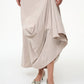A person wearing the Staud Viola Dress, a pale beige draped style with an asymmetrical hem, paired with pointed snakeskin-patterned shoes, stands against a plain white background.