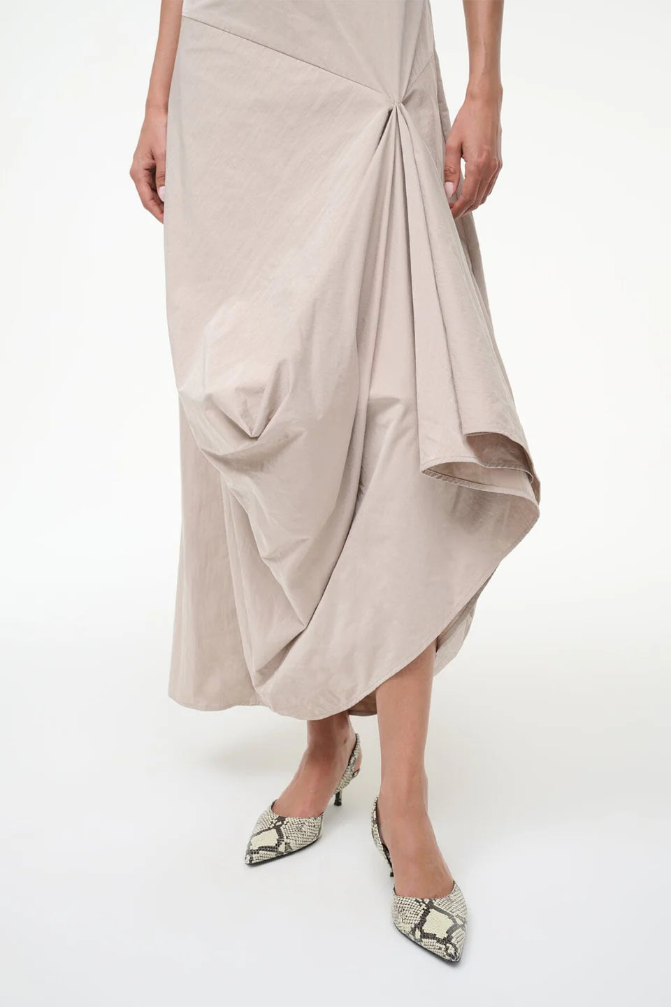 A person wearing the Staud Viola Dress, a pale beige draped style with an asymmetrical hem, paired with pointed snakeskin-patterned shoes, stands against a plain white background.