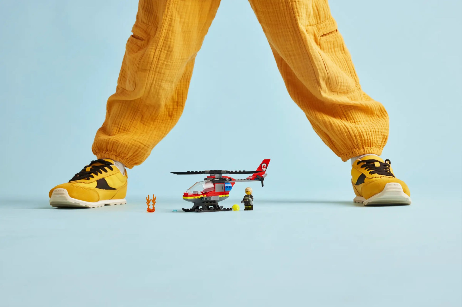 A person in yellow pants and sneakers stands with legs apart behind the LEGO® City Fire Rescue Helicopter by Legos - Toyhouse, a fireman figurine, and toy fire accessories on a light blue background.