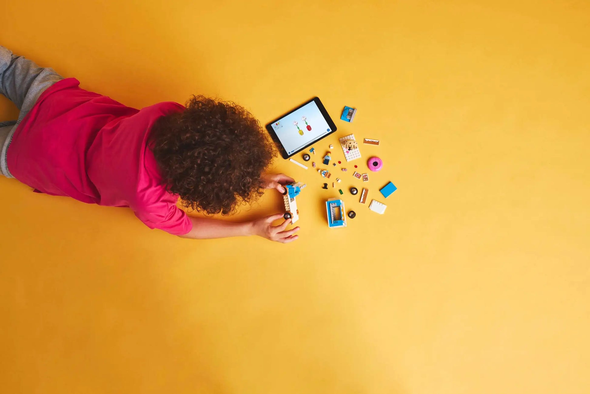 A child arranges vehicle toy parts on a yellow floor near a tablet showing images of the LEGO® City Donut Truck by Legos - Toyhouse.