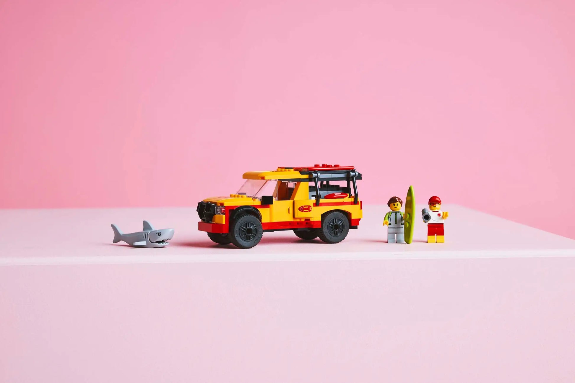 A yellow and red LEGO® City Lifeguard Beach Rescue Truck by Legos - Toyhouse, two toy figures with a surfboard, and a toy shark are arranged in a row against a pink background.