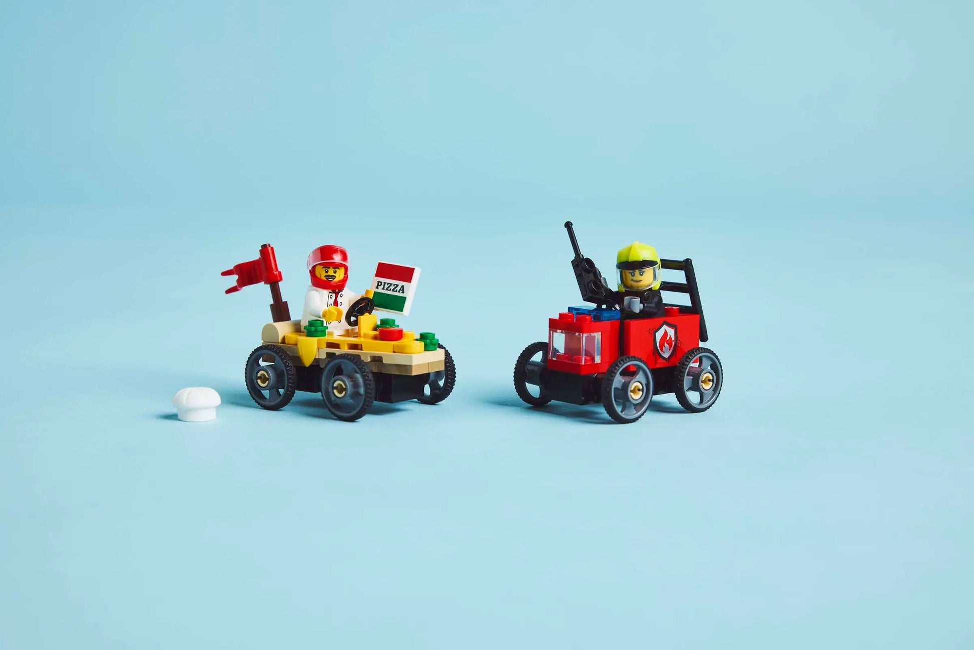 Two race cars from the Legos - Toyhouse LEGO® City Pizza vs. Fire Truck Race Car Pack speed across a blue backdrop—one with a pizza flag and red-helmeted driver, the other with a yellow-helmeted driver and special accessory.
