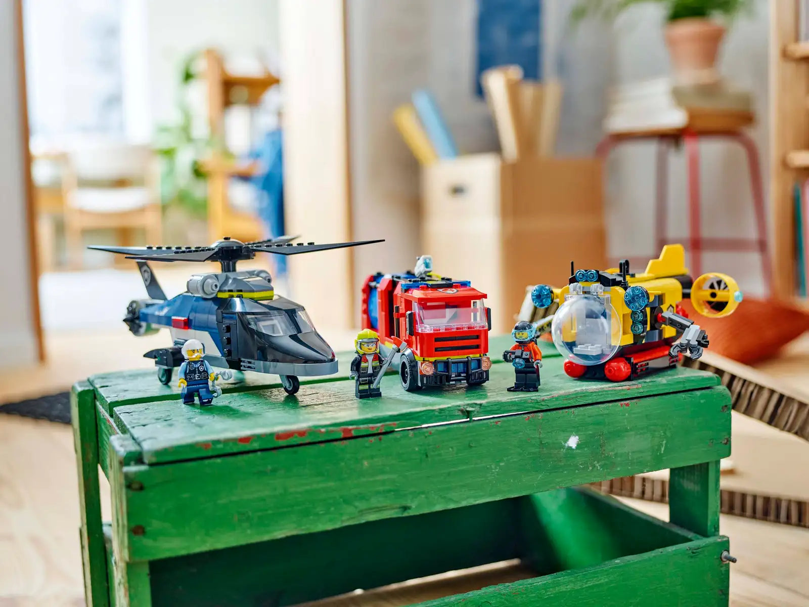 Three vehicles from the Legos - Toyhouse LEGO® City Helicopter, Fire Truck & Submarine Remix set are shown on a green wooden crate, surrounded by several LEGO minifigures.