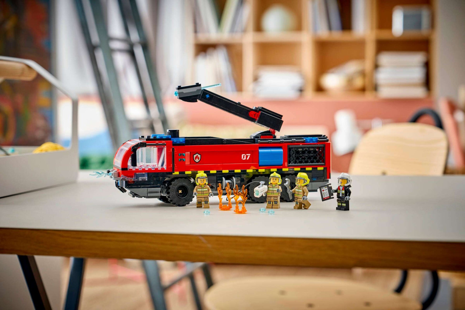 A Legos - Toyhouse LEGO® City Airport Fire Truck with an extending arm is on a table, surrounded by five firefighter minifigures and fire accessories in a brightly lit room.