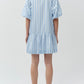 A person faces away, wearing the Simkhai Crissy Stripe Dress—a light blue and white vertically striped mini shirtdress with puff sleeves and a ruffled hem—paired with white shoes.