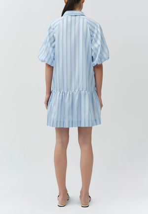 A person faces away, wearing the Simkhai Crissy Stripe Dress—a light blue and white vertically striped mini shirtdress with puff sleeves and a ruffled hem—paired with white shoes.