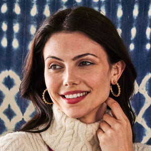 Woman with long dark hair in a white turtleneck and Juliska Capucine De Wulf Berry Icon Medium Hoop Earrings smiles while touching her earring, set against a blue and white patterned background.