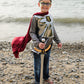 A child dressed in the Great Pretenders Silver Knight with Cape, 5-6Y stands barefoot on a rocky beach, wearing a crown and smiling at the camera while holding a toy sword.
