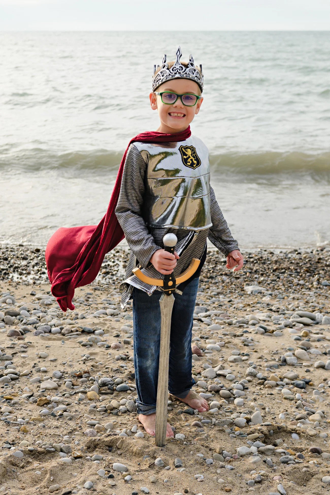 A child dressed in the Great Pretenders Silver Knight with Cape, 5-6Y stands barefoot on a rocky beach, wearing a crown and smiling at the camera while holding a toy sword.