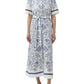 A person wears the Simkhai Audria Linen Midi Dress in white and blue with a floral and seashell print, short sleeves, belted waist, and button-down front, paired with white heeled sandals.