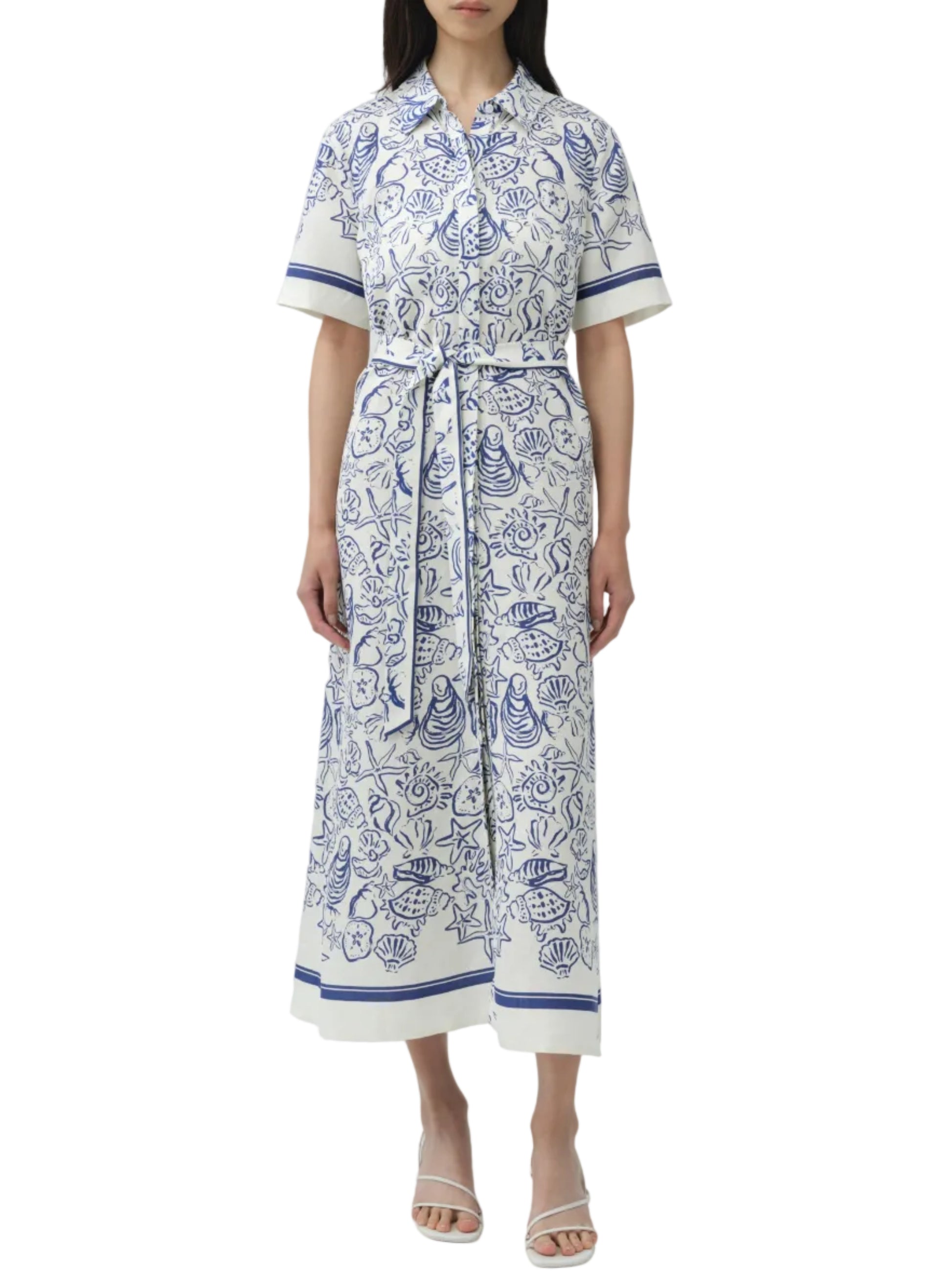 A person wears the Simkhai Audria Linen Midi Dress in white and blue with a floral and seashell print, short sleeves, belted waist, and button-down front, paired with white heeled sandals.