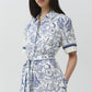 A woman wears the Simkhai Audria Linen Midi Dress, featuring short sleeves, a belted waist, and a blue and white seashell and starfish pattern, standing against a plain background.