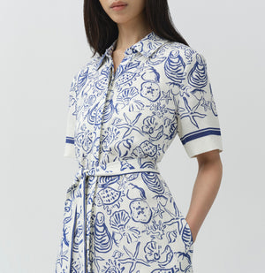 A woman wears the Simkhai Audria Linen Midi Dress, featuring short sleeves, a belted waist, and a blue and white seashell and starfish pattern, standing against a plain background.