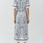 A woman stands with her back to the camera, wearing the Simkhai Audria Linen Midi Dress with blue seashell and starfish patterns, paired with white open-toe heels.