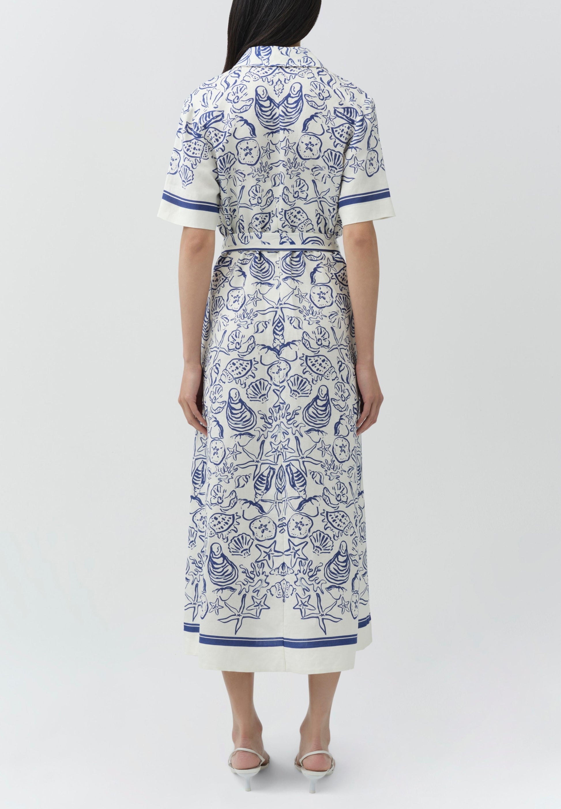 A woman stands with her back to the camera, wearing the Simkhai Audria Linen Midi Dress with blue seashell and starfish patterns, paired with white open-toe heels.