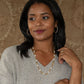 A woman stands before a textured brown background wearing a light gray sweater, gold hoop earrings, a Juliska Capucine De Wulf Berry & Bead Chain layered with a pearl and gold piece, and a gold ring.