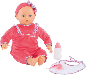 The Corolle Lila Chérie 17" baby doll, dressed in pink with a headband and made of soft-touch vinyl, sits beside a white bottle, pacifier, and matching bib.