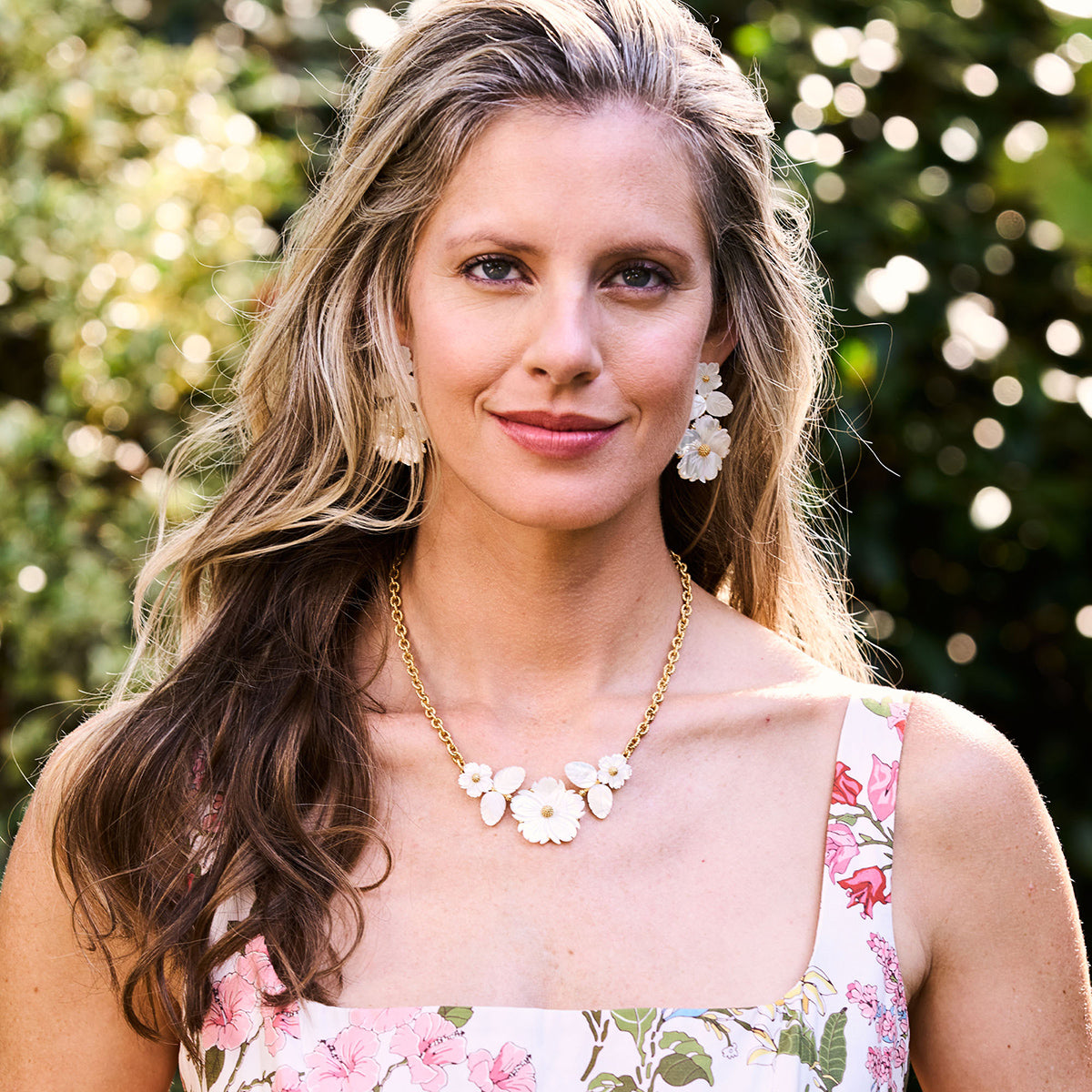 A woman with long, wavy blonde hair stands outdoors in greenery, wearing a floral dress, flower earrings, and the Mermaid Garden Bouquet Necklace by Juliska Capucine De Wulf.