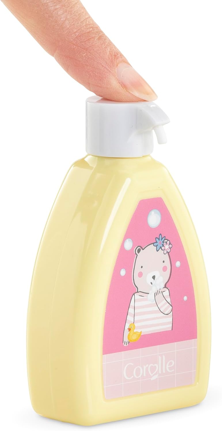 A hand presses the top of a yellow bottle with a pink label featuring a cartoon bear and "Corolle," part of the Corolle Floral Changing Accessories Set—ideal for your doll accessories or imaginative play on the go.