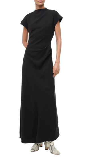 A person wearing the Staud Zaya Dress, a long black A-line maxi in triacetate crepe with a high neckline, paired with patterned ankle boots, stands against a white background.