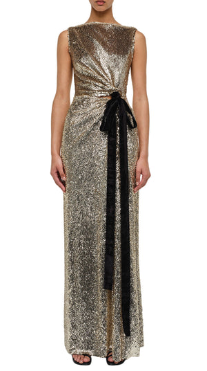 A woman wears the Simkhai Felicity Gown, a sleeveless metallic gold floor-length dress with liquid shimmer, side knot, and black velvet sash, paired with black open-toe heels.