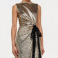 A person wearing the Simkhai Felicity Gown—a sleeveless, metallic gold paillette sequin design with a black velvet waist bow—shown from shoulders to knees.
