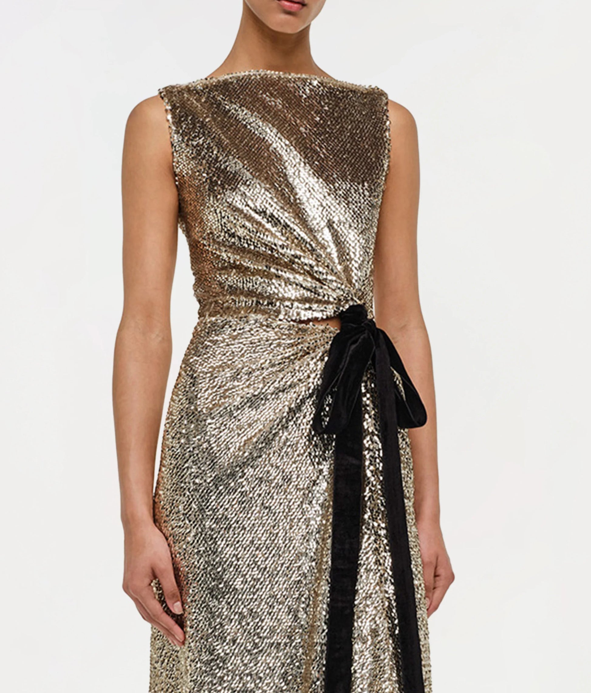 A person wearing the Simkhai Felicity Gown—a sleeveless, metallic gold paillette sequin design with a black velvet waist bow—shown from shoulders to knees.