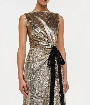 A person wearing the Simkhai Felicity Gown—a sleeveless, metallic gold paillette sequin design with a black velvet waist bow—shown from shoulders to knees.
