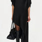 Wearing the Simkhai Sadia Cape Mini Dress with an asymmetric hemline and short sleeves, paired with a black handbag and knee-high pointed black boots, she exudes modern glamour against a plain background.