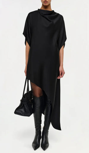 Wearing the Simkhai Sadia Cape Mini Dress with an asymmetric hemline and short sleeves, paired with a black handbag and knee-high pointed black boots, she exudes modern glamour against a plain background.