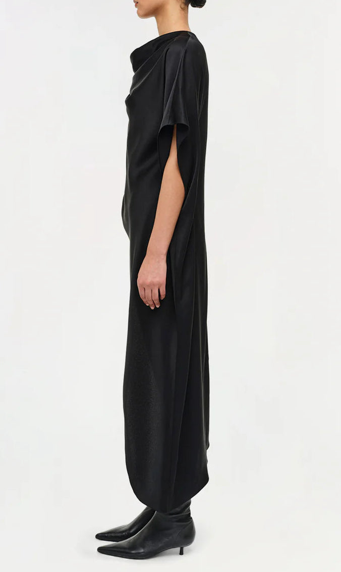 A person in profile models the Simkhai Sadia Cape Mini Dress—loose-fitting and black with short sleeves—paired with black pointed-toe boots, showcasing modern glamour against a plain white background.