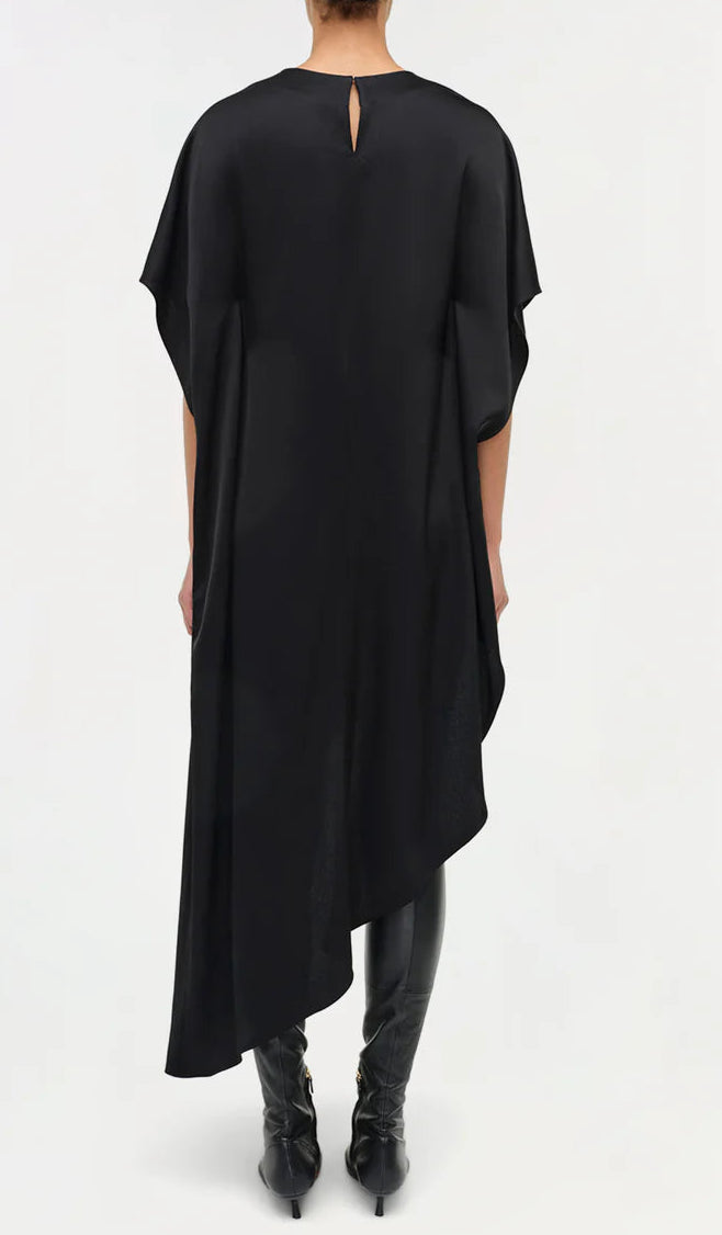 Shown from the back, a person wears the Simkhai Sadia Cape Mini Dress—black, knee-length with an asymmetric hemline and short sleeves—paired with black boots for an effortlessly modern look.