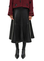Shown from shoulders to feet, a person in Simkhai's Ryma Leather Midi Skirt, styled with a burgundy jacket and black heeled boots, captures elevated dressing with a sleek silhouette.