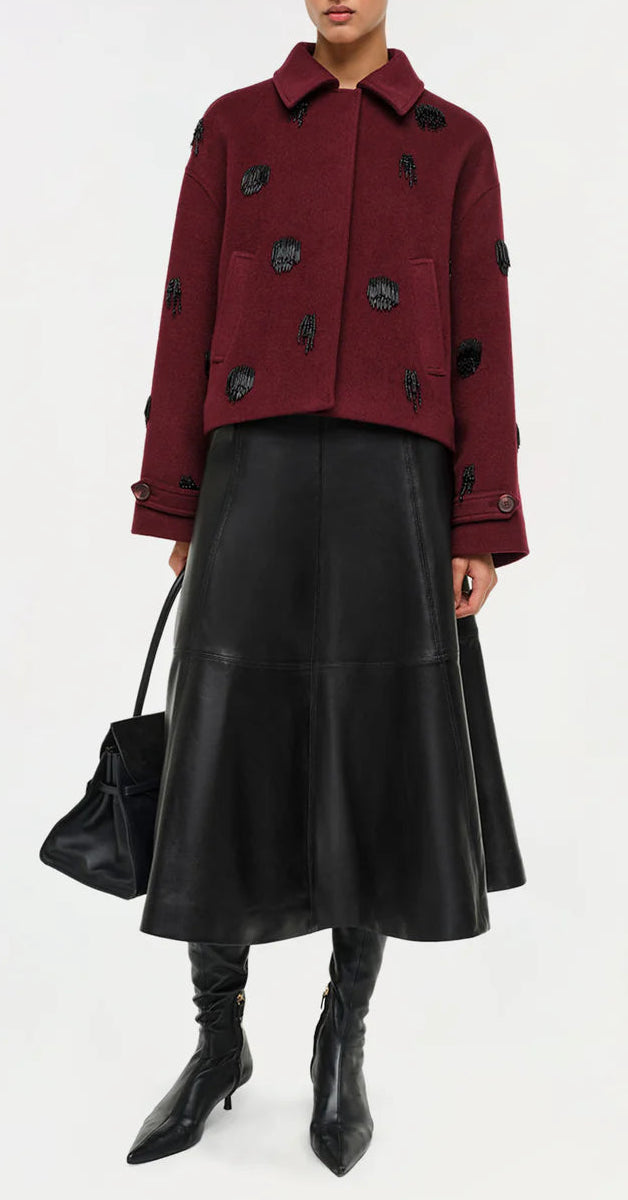 A person wearing a maroon jacket, black knee-high boots, a black handbag, and the Simkhai Ryma Leather Midi Skirt stands against a white background, showcasing a sleek silhouette ideal for elevated style.