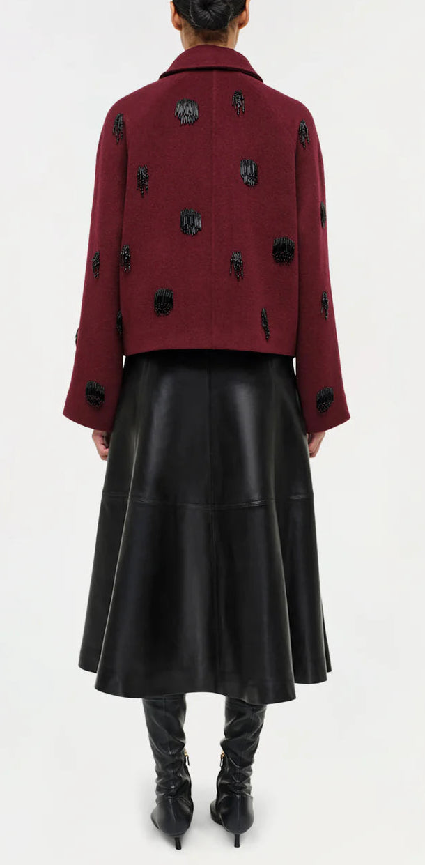 A person stands facing away, showcasing elevated style in a maroon jacket with black embellishments, the Simkhai Ryma Leather Midi Skirt, and knee-high black boots.