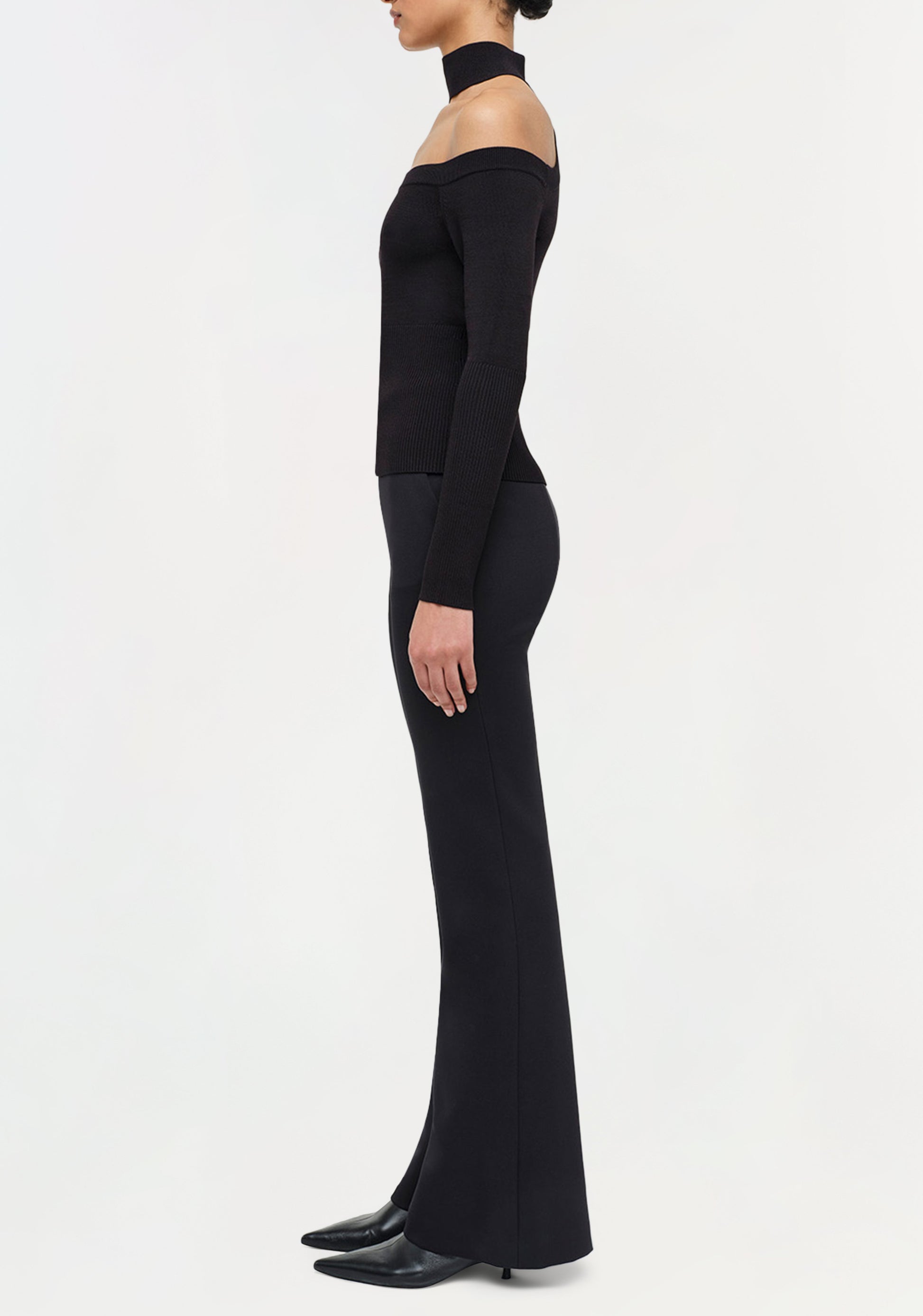A person stands in profile against a white background, wearing an off-the-shoulder black top, Simkhai's Kenna Trouser for refined minimalism, and black pointed shoes.