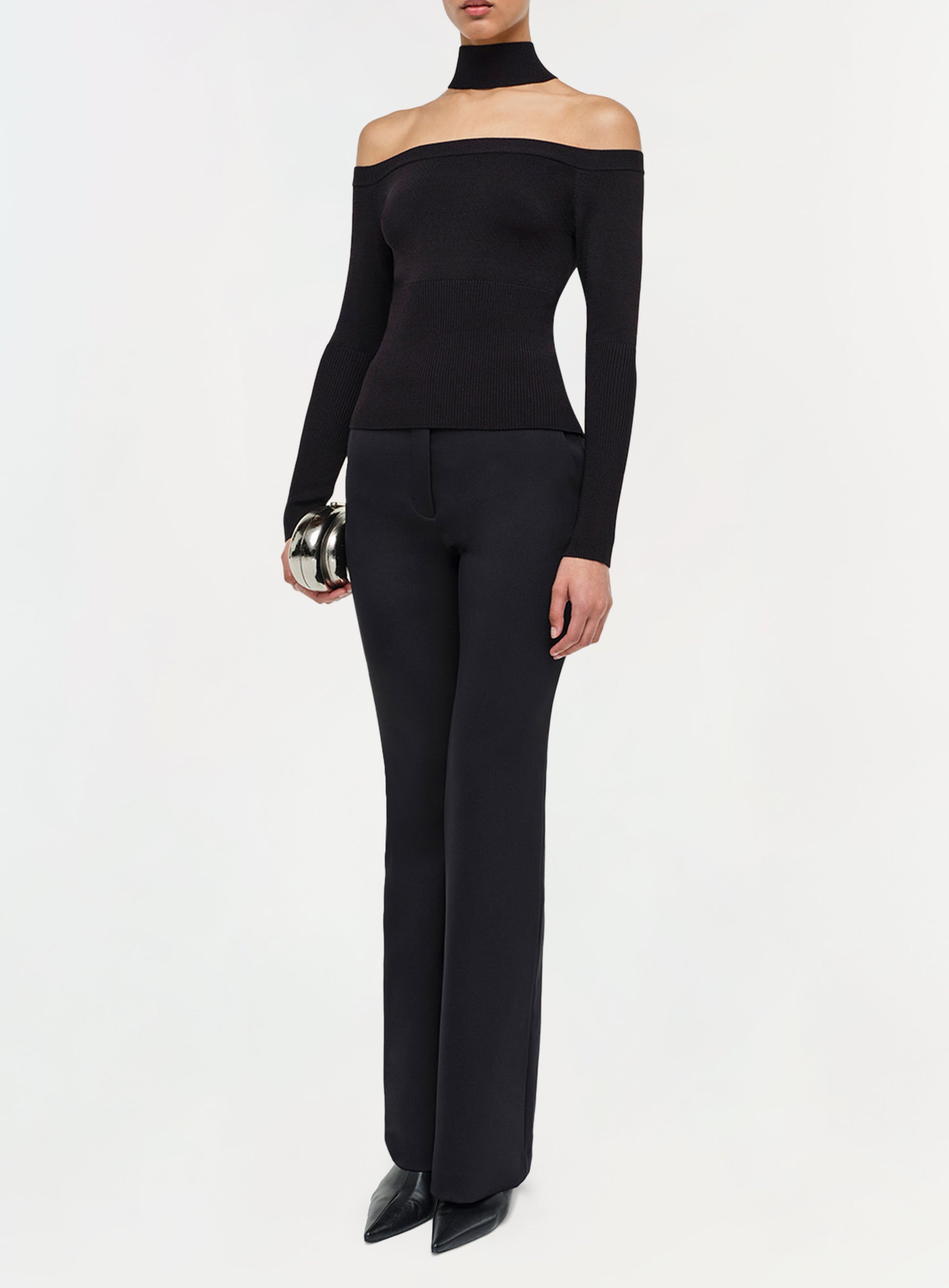 A person wears an off-the-shoulder black top, Simkhai Kenna Trousers with a flared silhouette, black shoes, and a choker, holding a small metallic clutch—an effortlessly refined minimalist look.