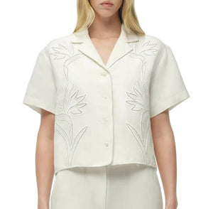 A woman wears the Simkhai Louisa Camp Shirt in off-white with floral embroidery, paired with matching pants for an effortlessly chic look.