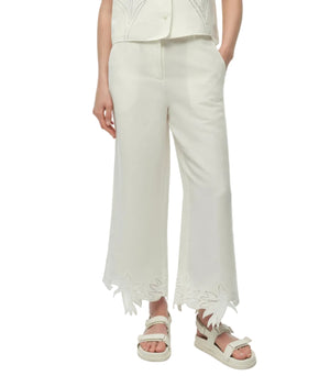 Model wears Simkhai's Kallin Cropped Wide Leg Pant, featuring a leaf-shaped cutout hem, paired with a white sleeveless top and sandals. Hands in pockets; upper body partially out of frame.