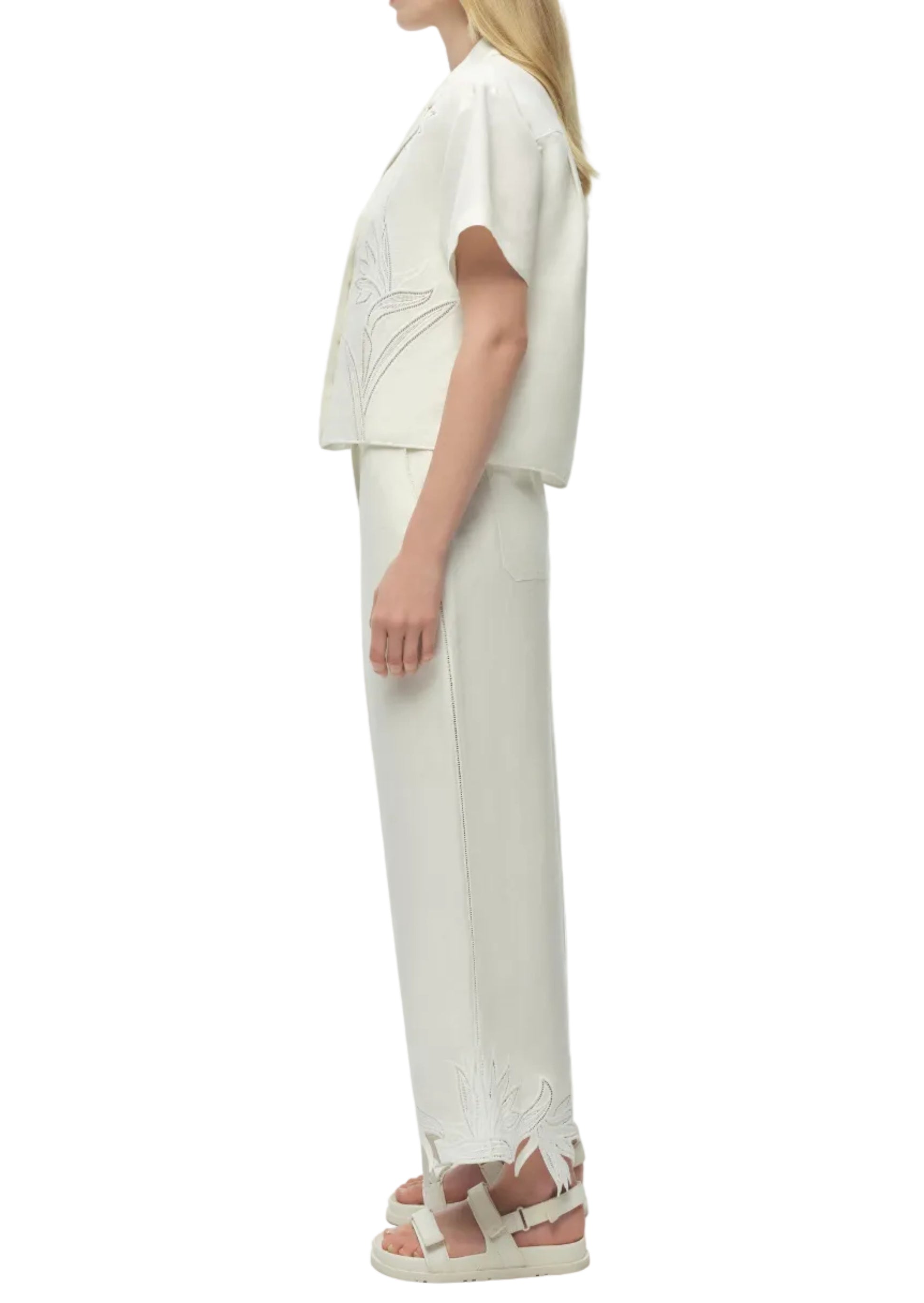 Woman in side profile wears the Simkhai Louisa Camp Shirt in white with floral embroidery, matching wide-leg pants, and white platform sandals.