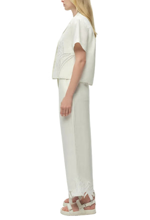 A woman stands in profile wearing a white short-sleeve top and Simkhai Kallin Cropped Wide Leg Pant featuring floral embroidery near the hem, paired with white sandals.